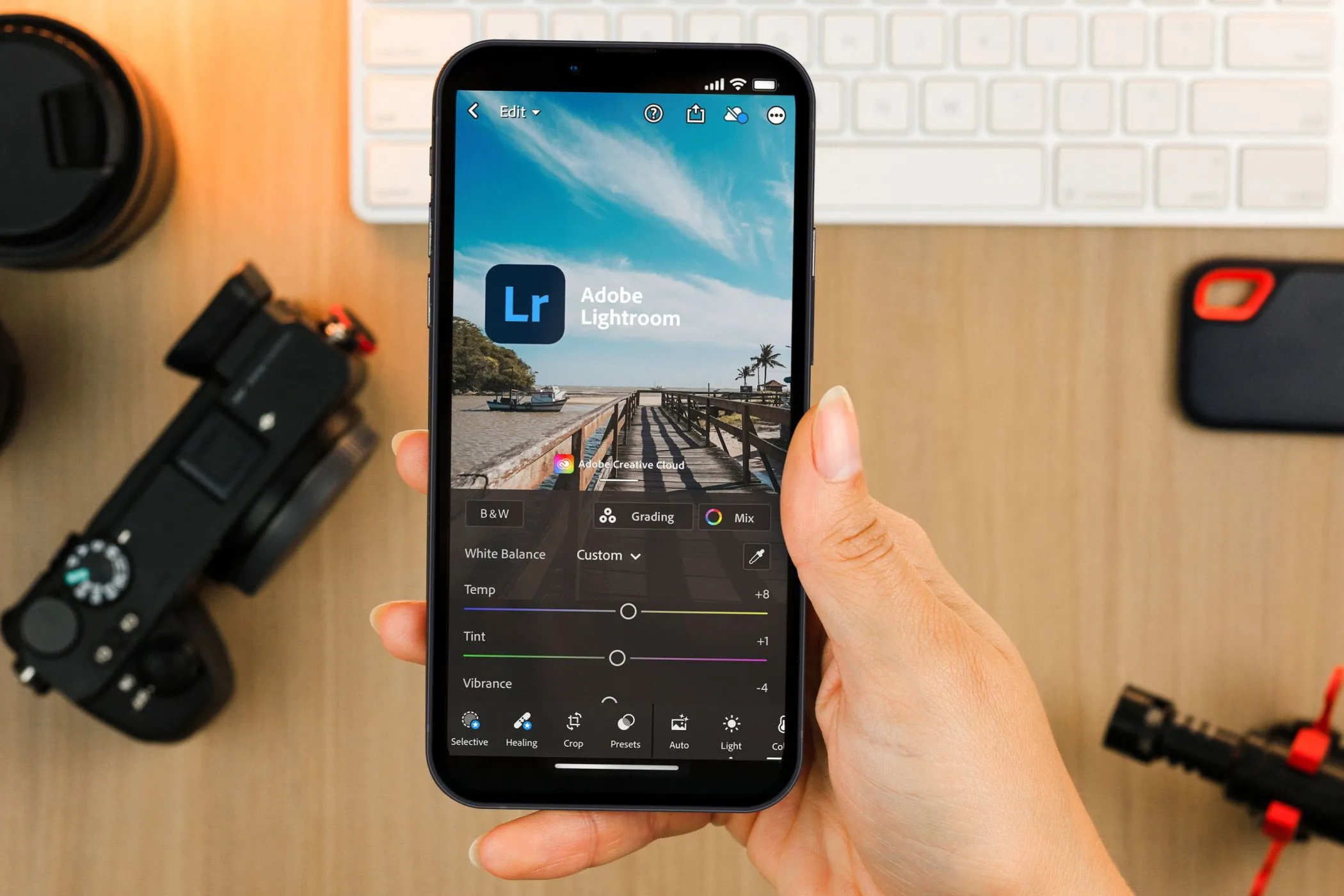 Bàn tay đang cầm điện thoại hiển thị ứng dụng Lightroom Mobile, phía sau là ống kính máy ảnh và phụ kiện
