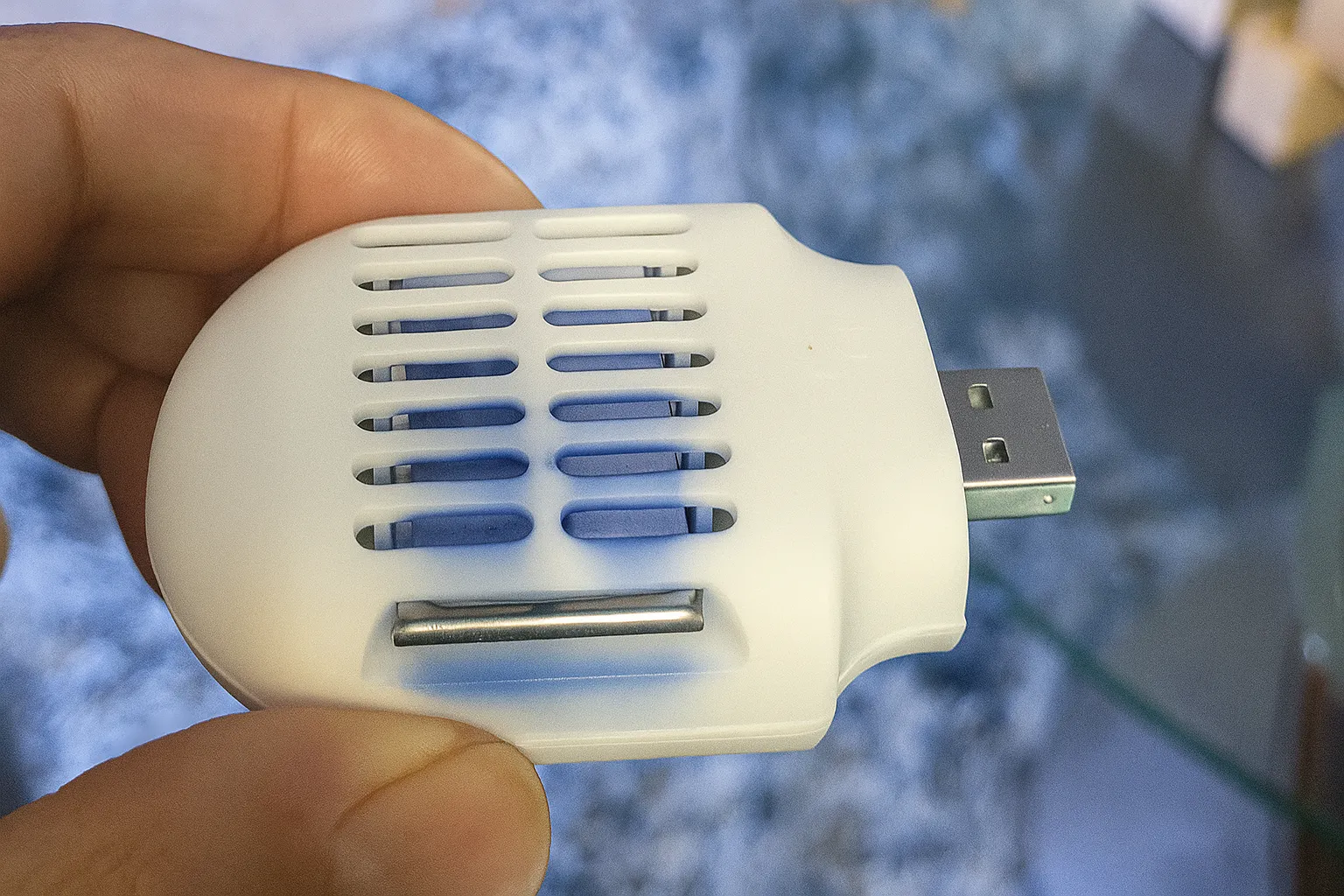 Bàn tay đang cầm thiết bị đuổi muỗi nhỏ gọn, màu trắng, cấp nguồn qua USB.