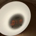 Đồng hồ báo thức giả lập bình minh đặt trên sàn, phát ra ánh sáng vàng dịu giúp thức dậy tự nhiên.