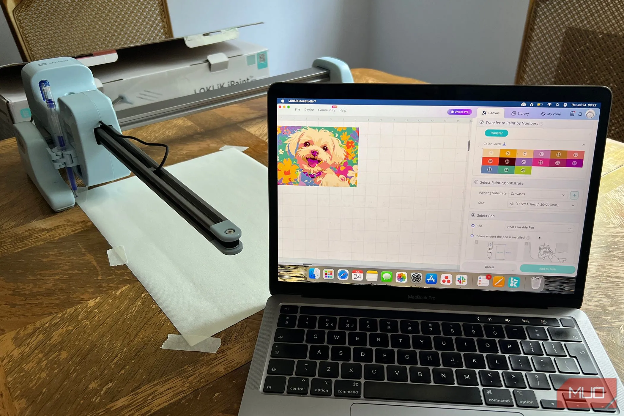 Máy LOKLiK iPaint đặt cạnh tấm canvas trống và ảnh gốc được hiển thị trên MacBook