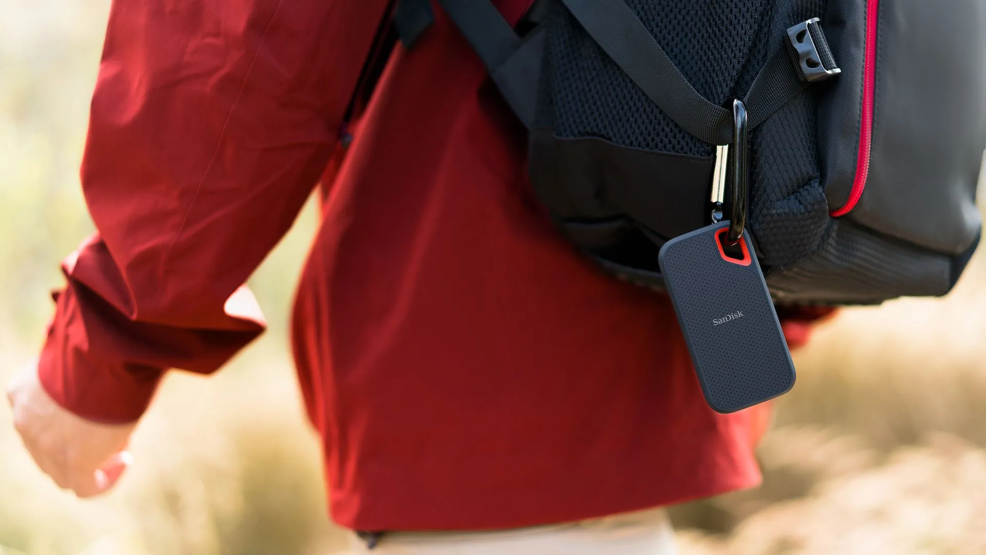 Người đàn ông mặc áo khoác đỏ đeo ba lô, móc ổ cứng SSD SanDisk Extreme Portable 8TB màu đen lên ba lô, thể hiện tính tiện lợi khi di chuyển.