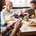 Người đàn ông trẻ đang sử dụng điện thoại thông minh tại quán cà phê, bên cạnh bạn bè, để truy cập Wi-Fi miễn phí.