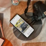 Người dùng đang trải nghiệm Photoshop trên điện thoại Android đặt trên giường cùng máy ảnh và sổ tay